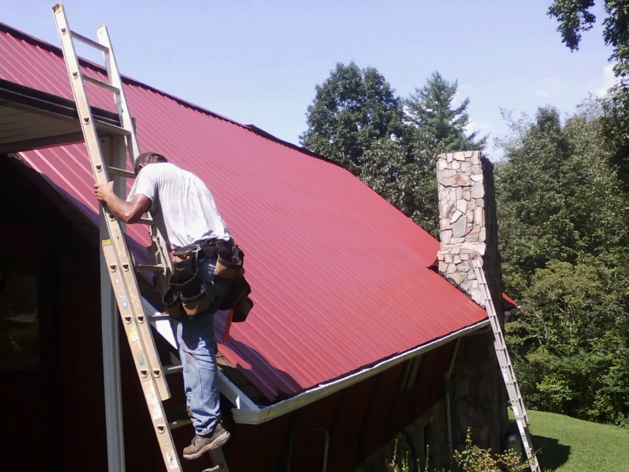 Roofer on a ladder beside a red metal roof and stone chimney — Soffit & Fascia Repair by Roofing Brothers in Santa Fe Springs